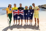 A group of Australian tennis players stand on a beach, some holding racquets, with an Australian flag in front of them.  
