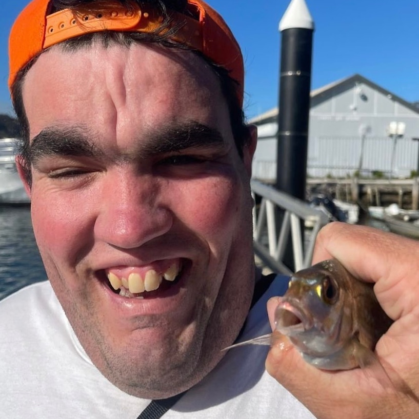 A young man with a backwards orange hat smiles while he holds a fish next to his face.