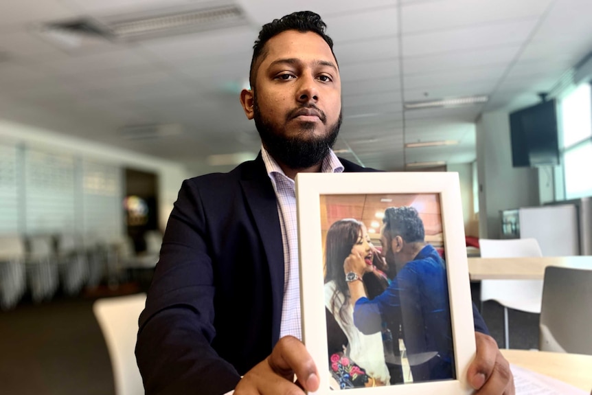 A Bangladeshi man in a suit holds a framed photograph of him and his wife.