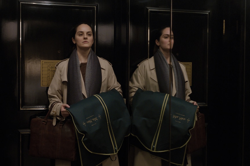 A brown-haired, 30-something woman stands next to a mirror in a lift, looking upset but defiant