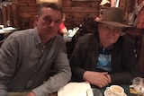 two men sit side by side at a dining table, the older one in an Akubra 