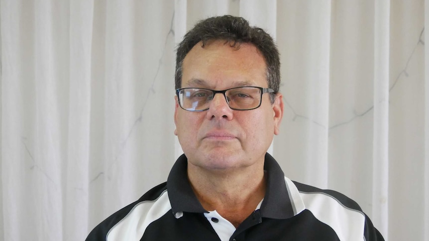 A man wearing a black shirt with white accents looks at the camera. He is wearing glasses and has dark hair.
