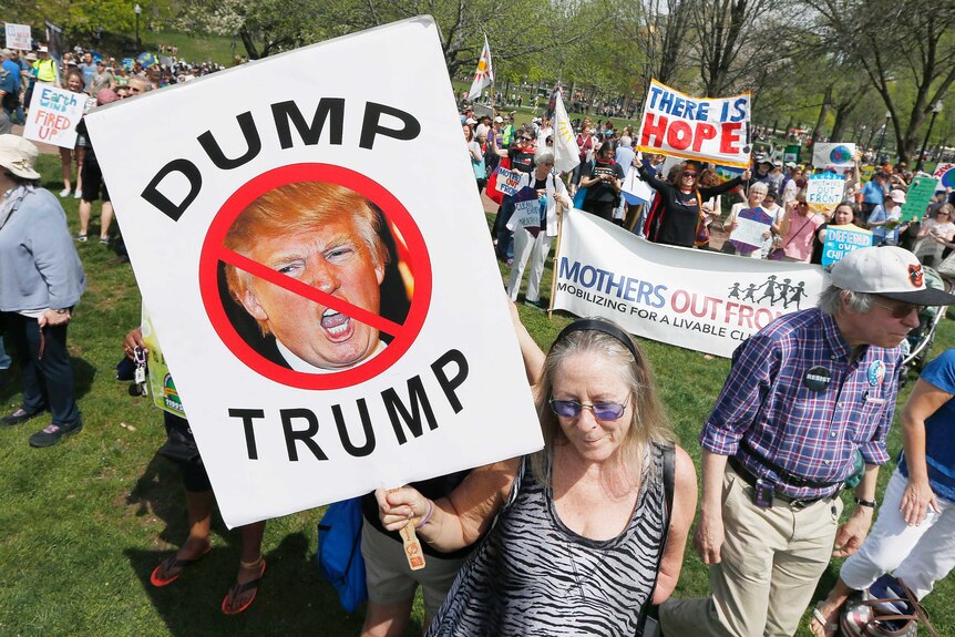 A woman holds a sign in her right hand reading "dump trump" with a photo of trump's face crossed out on it