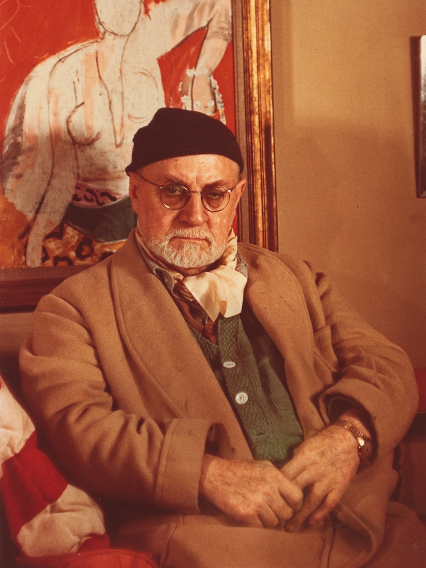 Colour photograph of the artist aged 79, wearing black knitted cap, camel coat and glasses, with beard, sitting in armchair.