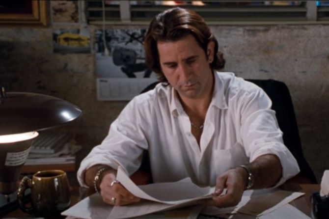 Anthony LaPaglia sits behind a desk in a white shirt reading a document