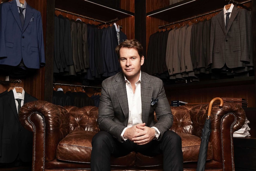 Fashion designer Matt Jensen, wearing a suit, sits on a chair in front of several racks of suits.