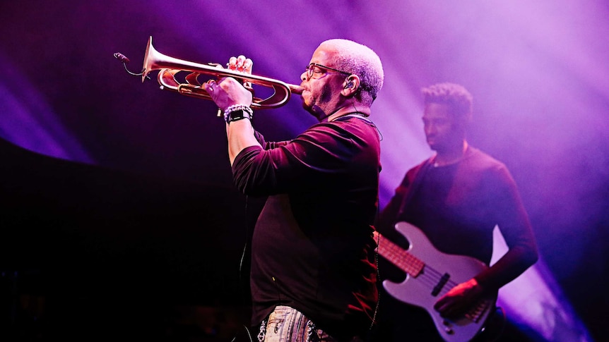Side profile photo of Terence playing trumpet on stage in front of a purple hazy background. A guitarist is standing behind him.