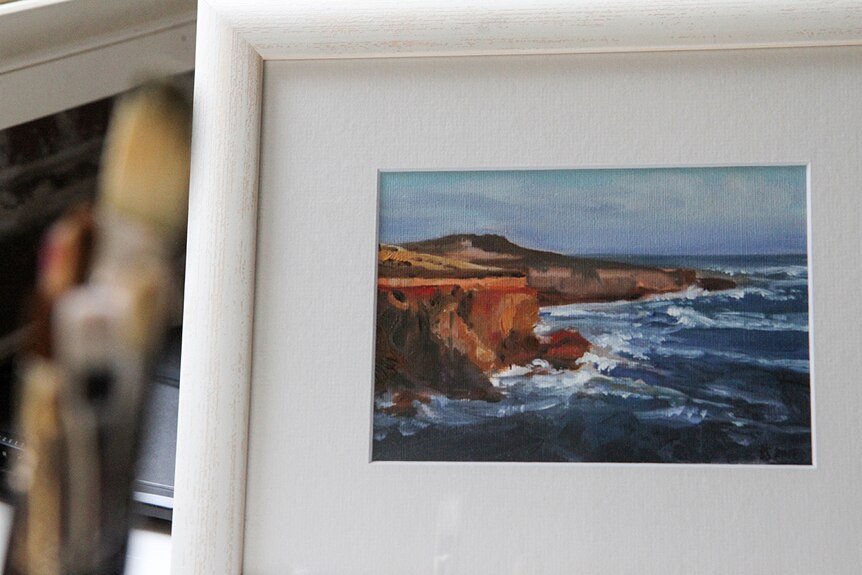 An artwork in a white frame, depicting a colourful image of rocky, red coastline being lapped by a deep blue ocean.