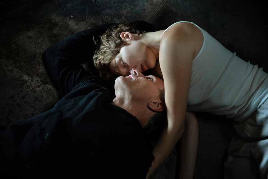 Blonde white woman in singlet lies intimately entwined with white man in black jumper; their faces are touching.