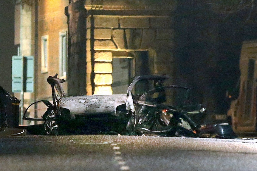 The charred wreckage of a car has collapsed into a twisted pile of metal and glass in the middle of a road.