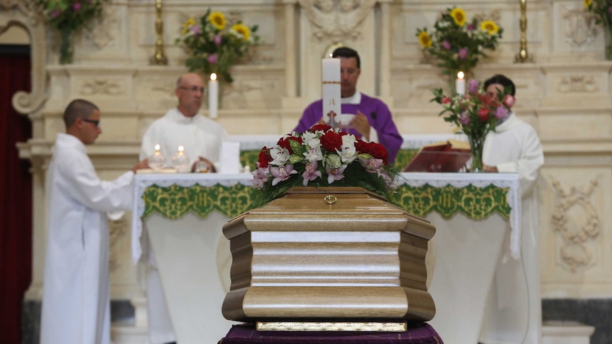 A coffin sits inside a church during a funeral, with a priest and other religious officials in the background.