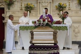 A coffin sits inside a church during a funeral, with a priest and other religious officials in the background.