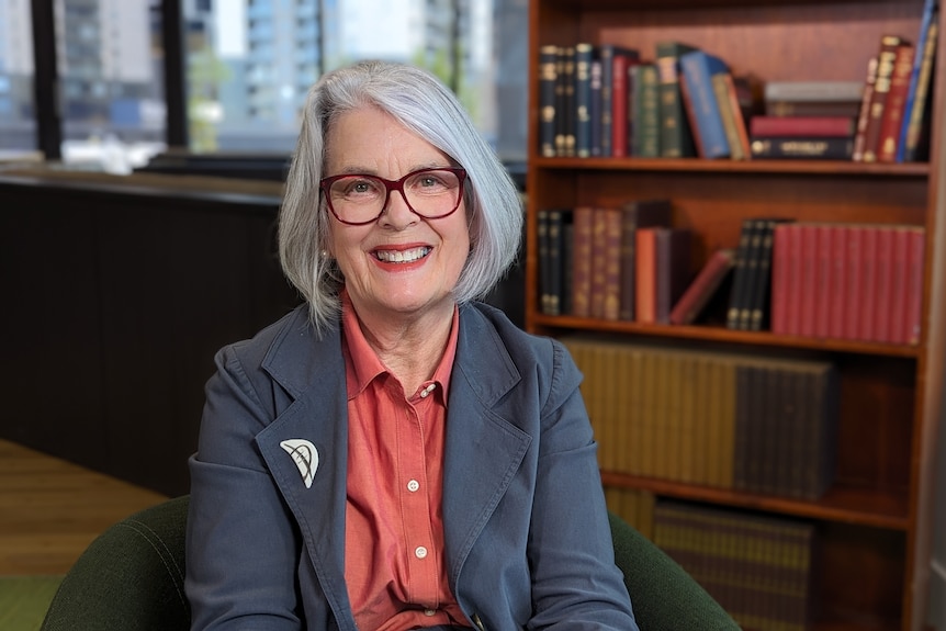 A women wearing a jacket and glasses smiles at the camera, as she sits in an office with a bookshelf in the background.