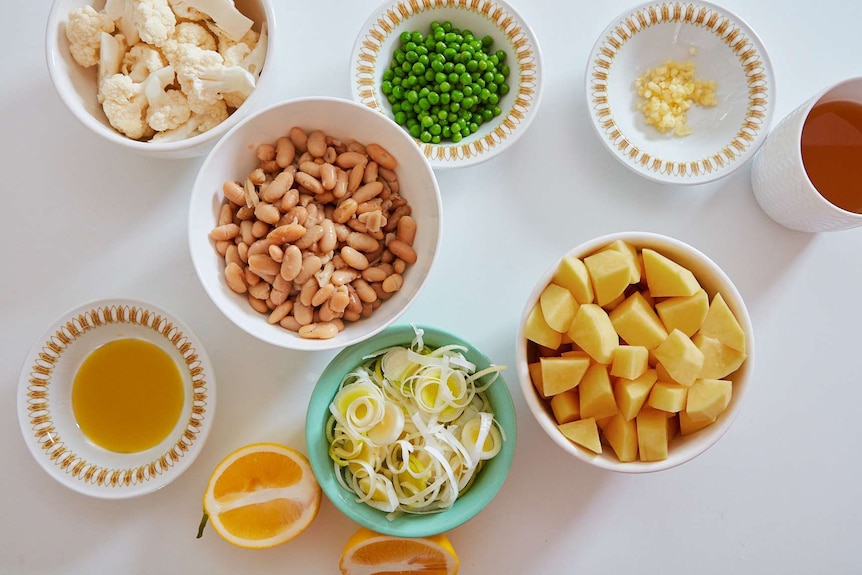 Soup ingredients laid out in bowls including cauliflower, cannellini beans, peas, chopped leaks, garlic, potatoes and lemon.