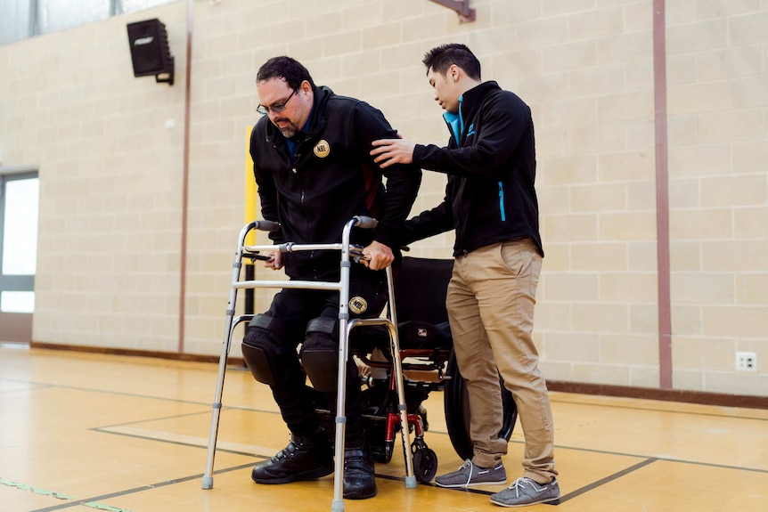 A man gets out of a wheelchair holding onto a walking frame with help from a physiotherapist.