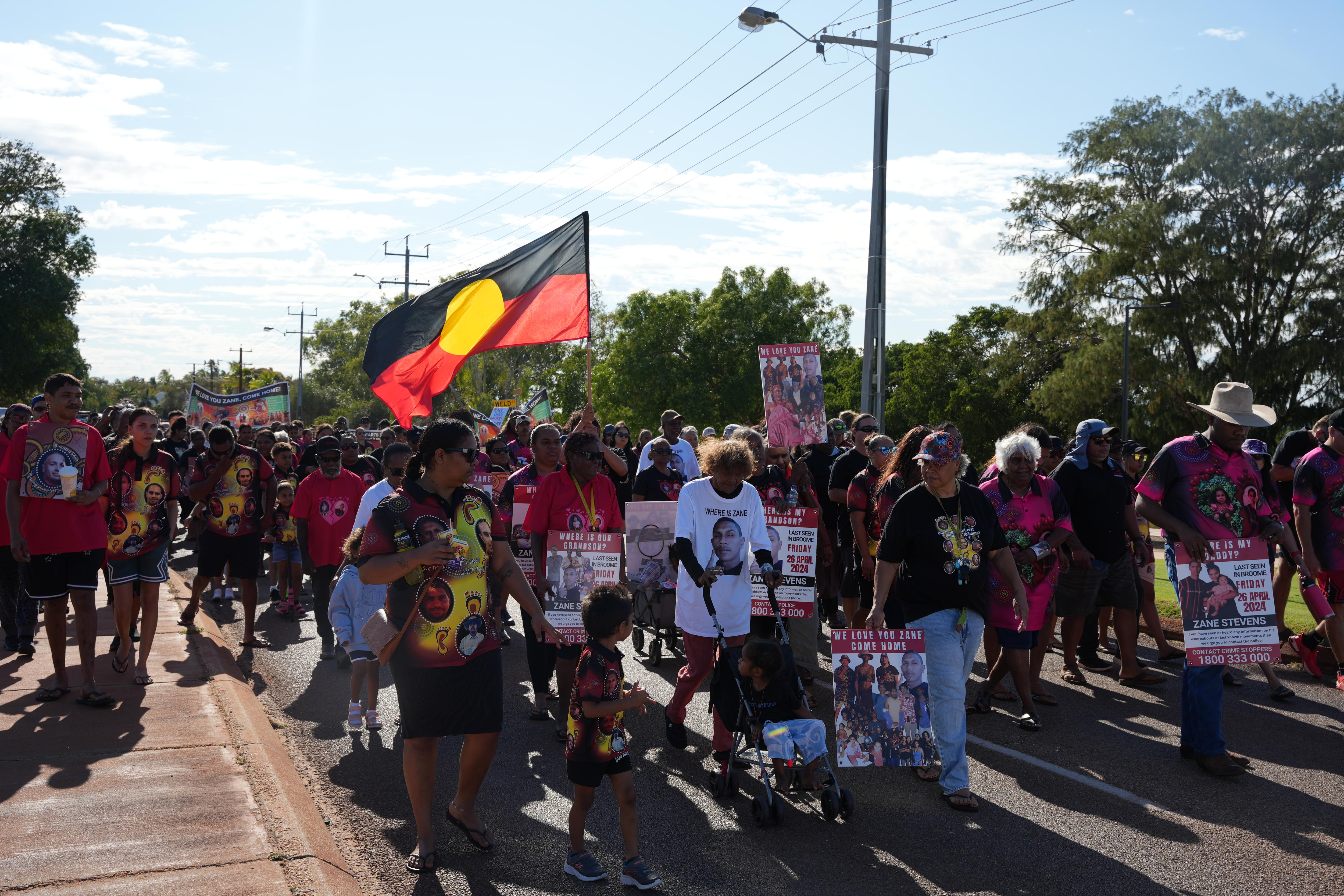 A large group of people march down a street. One holds an Aboriginal flag.