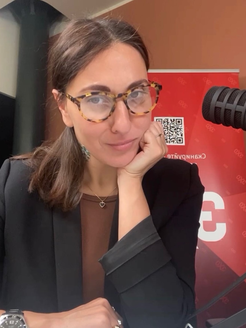 A young woman wearing tortoise shell glasses and black blazer leans her chin on her hand, next to a radio microphone