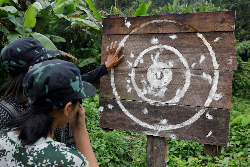 Two women wearing caps look at a wooden target in a green jungle.