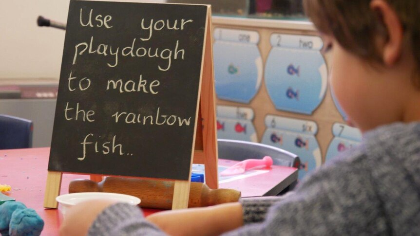 A small blackboard sits on a table in a classroom with the message, "Use your playdough to make the rainbow fish..".