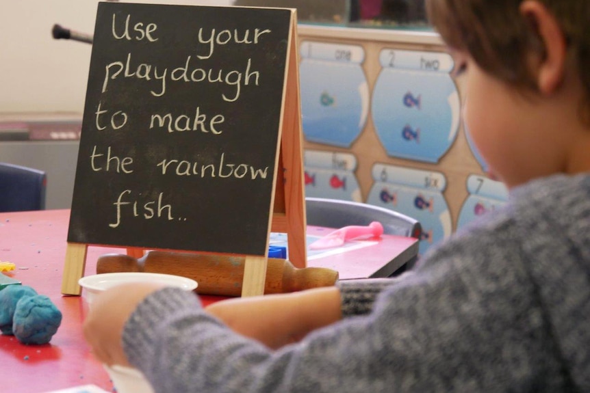 A small blackboard sits on a table in a classroom with the message, "Use your playdough to make the rainbow fish..".