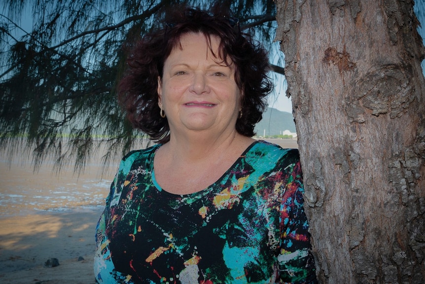 A woman stands next to a tree, smiling at the camera. She is wearing a patterned top