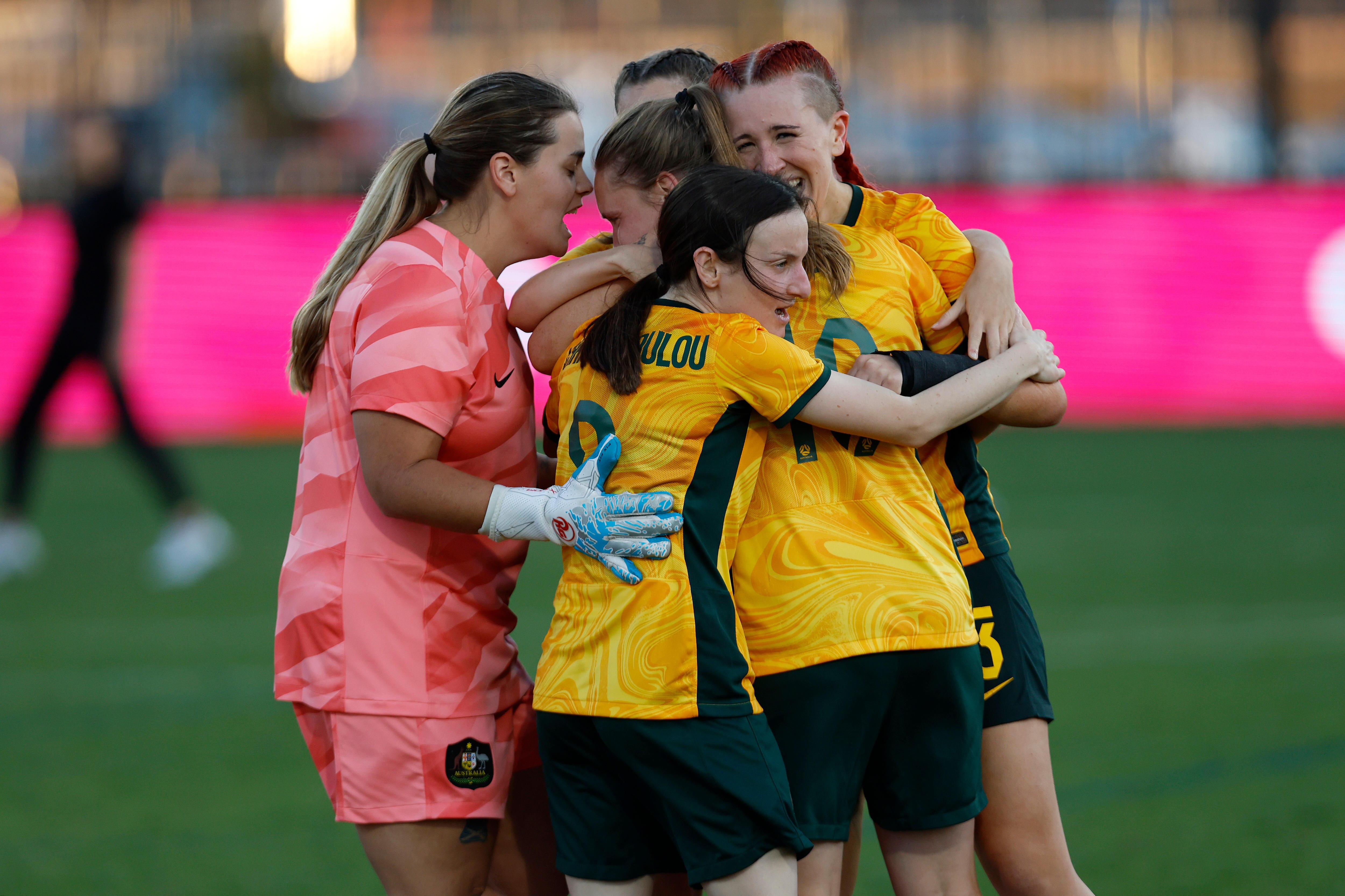 Four women in green and gold Australian jerseys and one in a pink gk jersey huddle in celebration on a soccer field.
