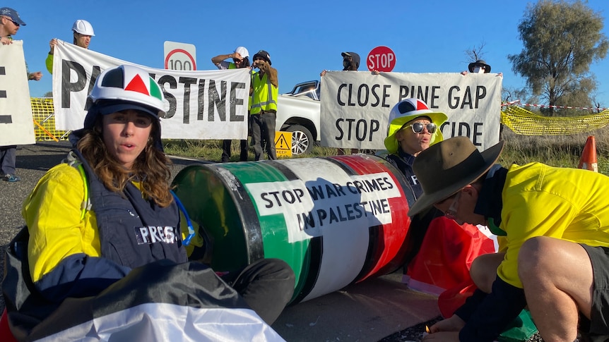 A group of people block the road with pro-Palestine signs. Three people are sitting on the road by an oil drum.