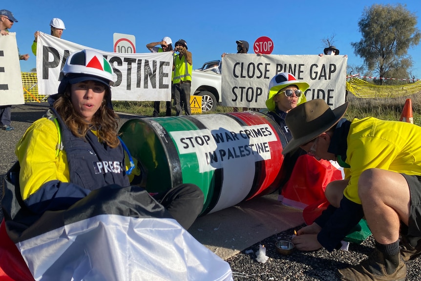 A group of people block the road with pro-Palestine signs. Three people are sitting on the road by an oil drum.