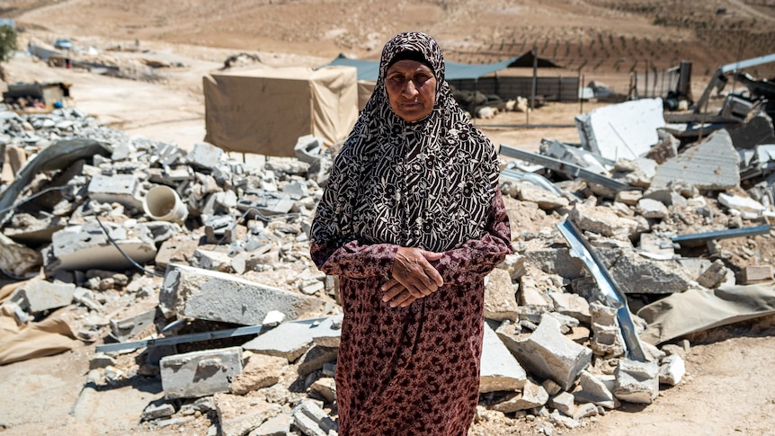 A woman in red patterned long dress and floral printed head covering stands in front of a pile of rubble