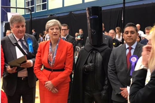 A man wearing a long black column on his head and a cape stands next to Prime Minister Theresa May