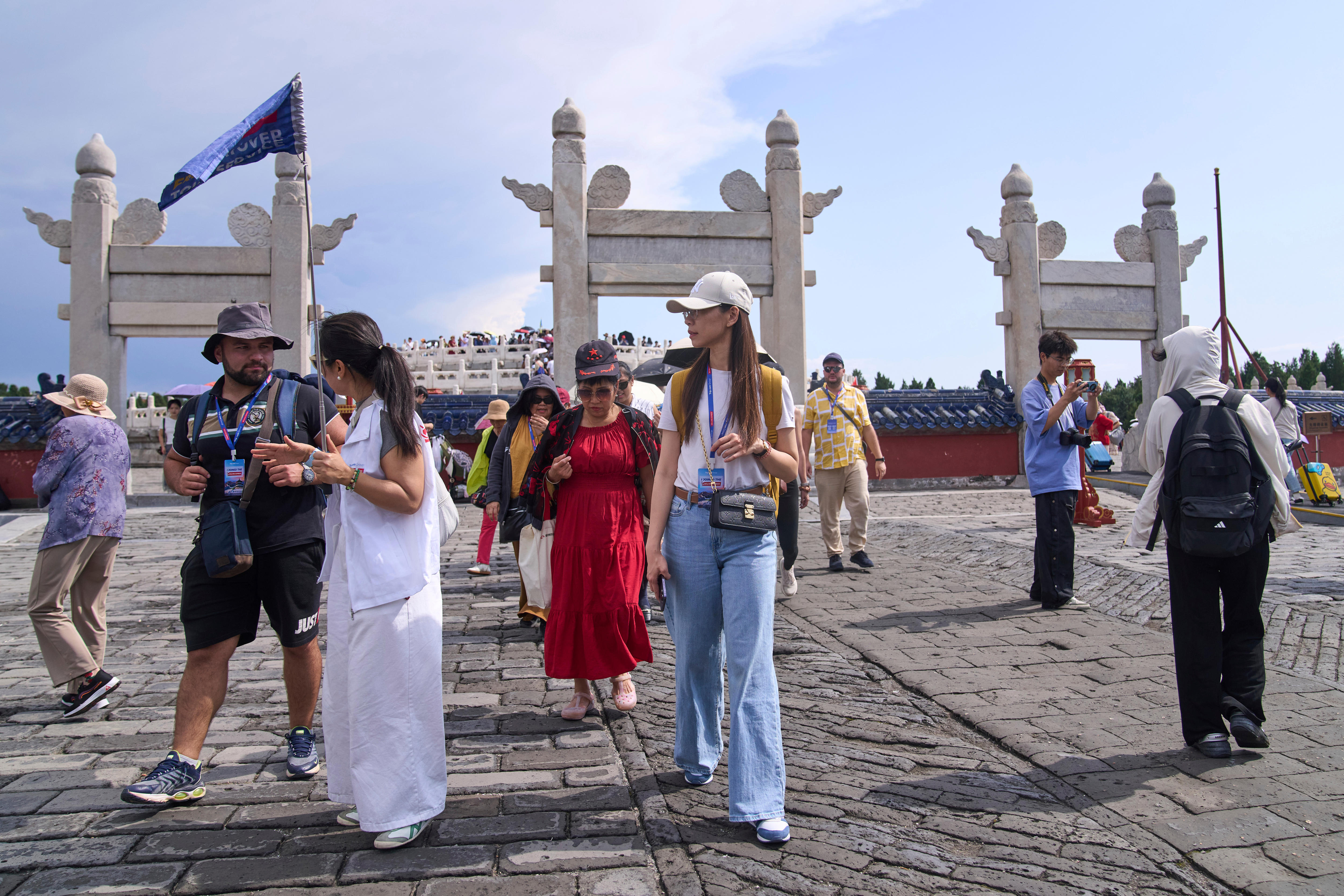 woman with long hair, jeans, baseball cap walks as part of tour group, while tour guide in white shirt and pants talks to man