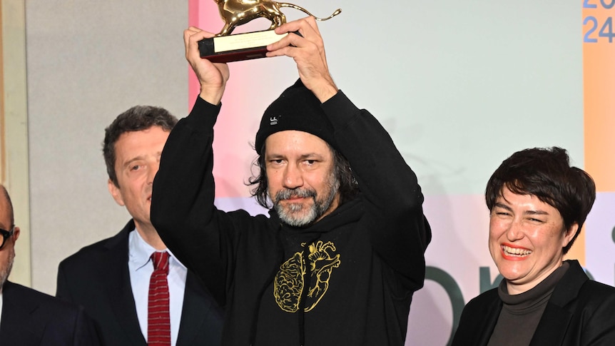 A Bigambul and Kamilaroi artist wearing a black hoodie holds up a golden lion trophy onstafe with thre other people.