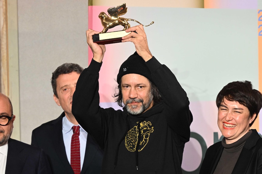 A Bigambul and Kamilaroi artist wearing a black hoodie holds up a golden lion trophy onstafe with thre other people.