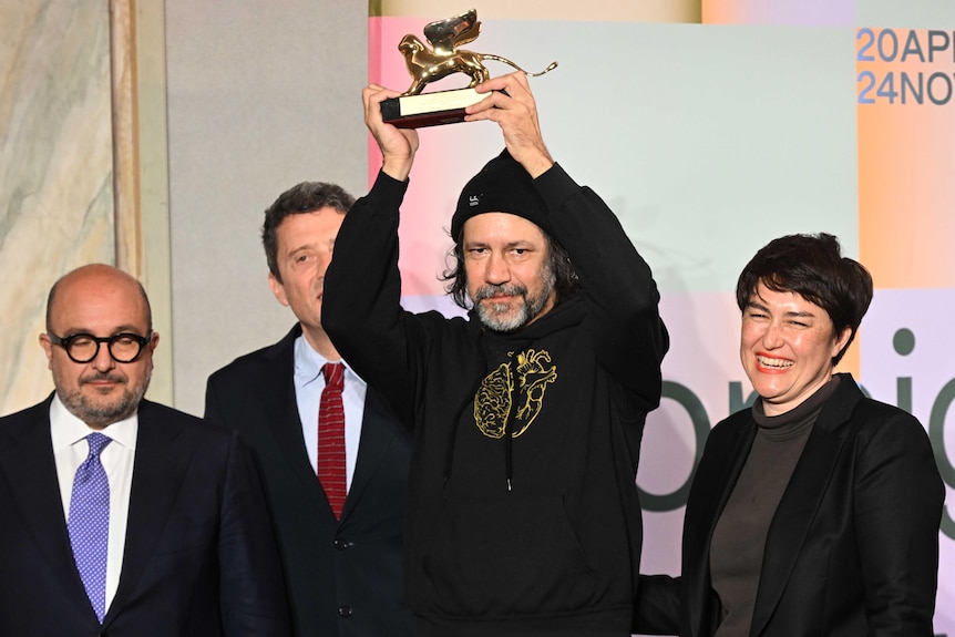 A Bigambul and Kamilaroi artist wearing a black hoodie holds up a golden lion trophy onstafe with thre other people.