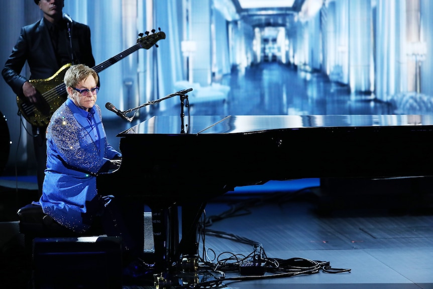 Elton John sits at a grand piano in a blue, sparkly suit, against a blue background.