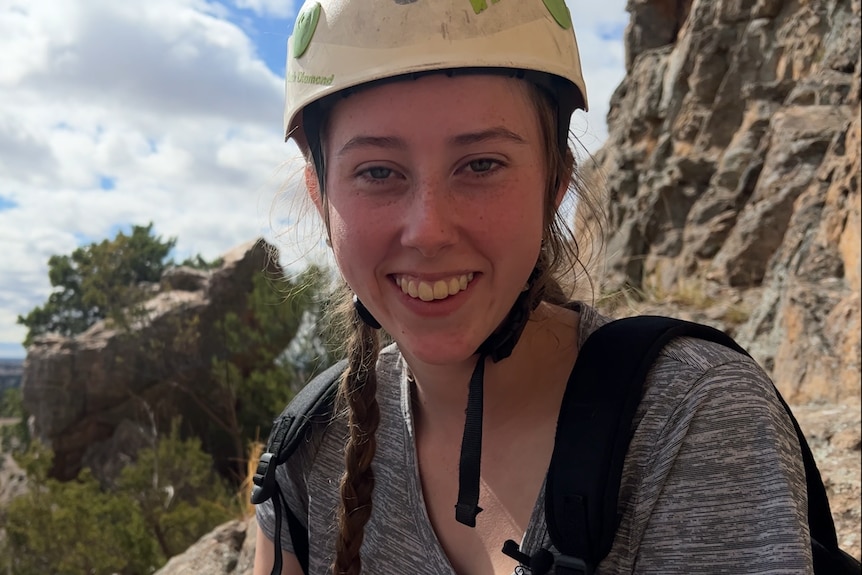 a girl with blond hair in braids, wearing a white helmet and grey tshirt sits at the base of a rocky mountain