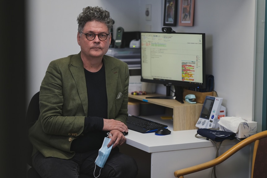 A man sits at an office desk holding a mask.