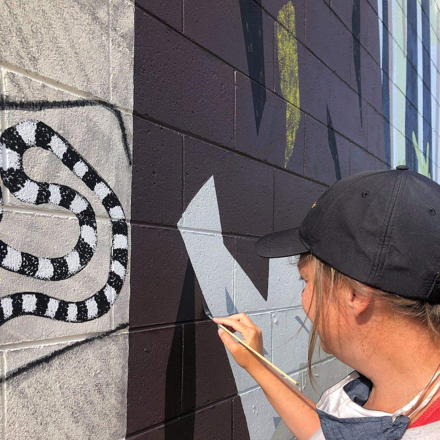 Profile of woman's head in black cap, her hand holds up a paint brush to a colourfully painted brick wall