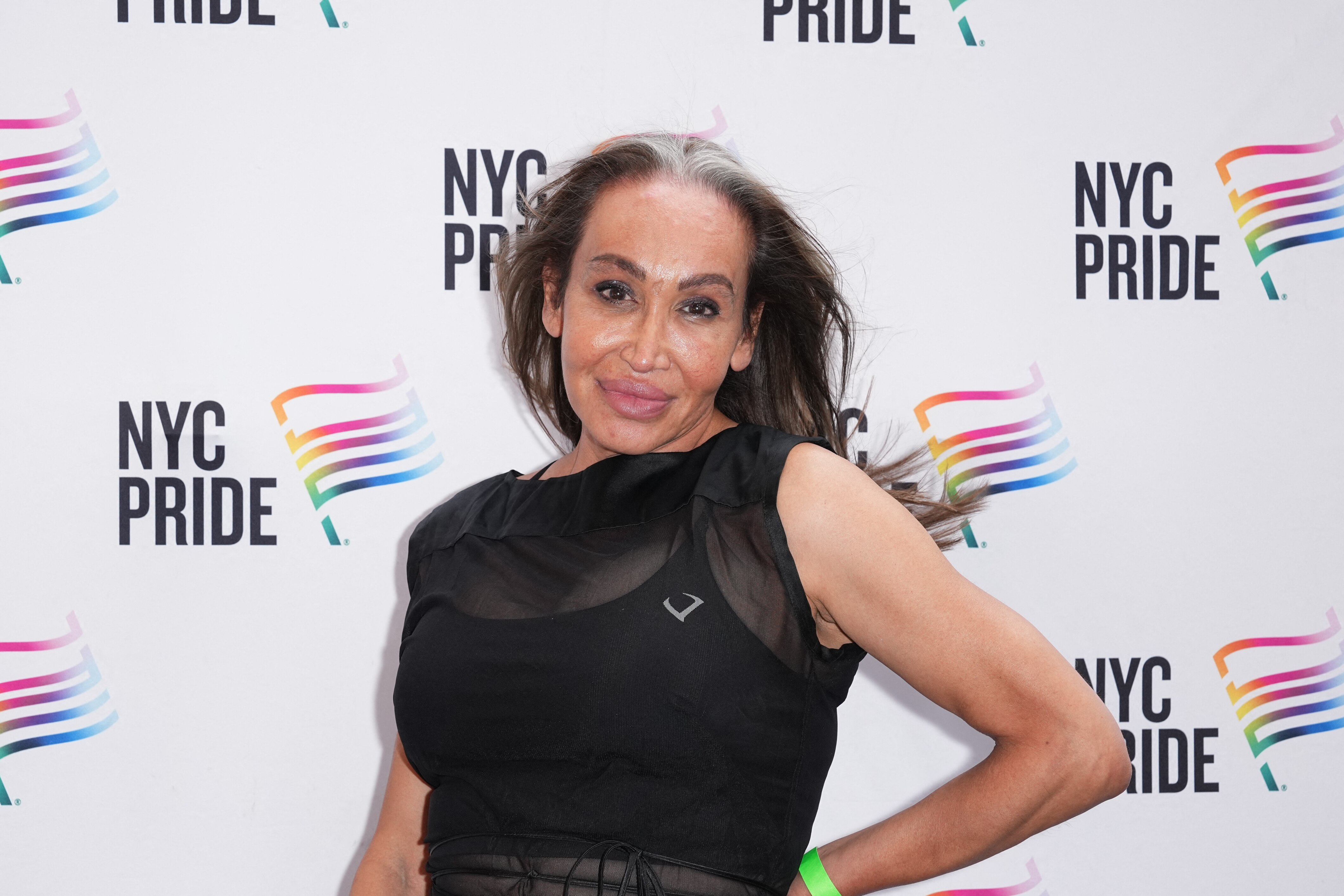 A close up of a woman with long hair standing and smiling in front of a wall with 'NYC Pride' logos all over it