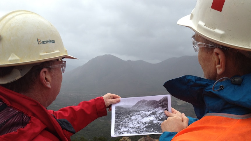 Two men hold an old photo of  Queenstown's Mount Lyell in front of the current scene