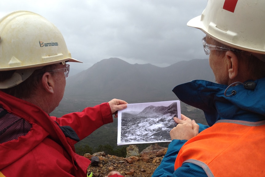 Two men hold an old photo of  Queenstown's Mount Lyell in front of the current scene