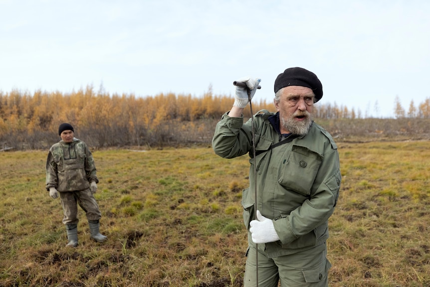 Sergey Zimov wearing white gloves holds a tool while standing in Pleistocene Park with a man behind him