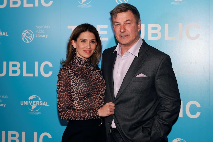 Hilaria and Alec Baldwin pose for cameras in front of a press wall, both wearing formal attire.