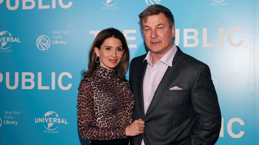 Hilaria and Alec Baldwin pose for cameras in front of a press wall, both wearing formal attire.