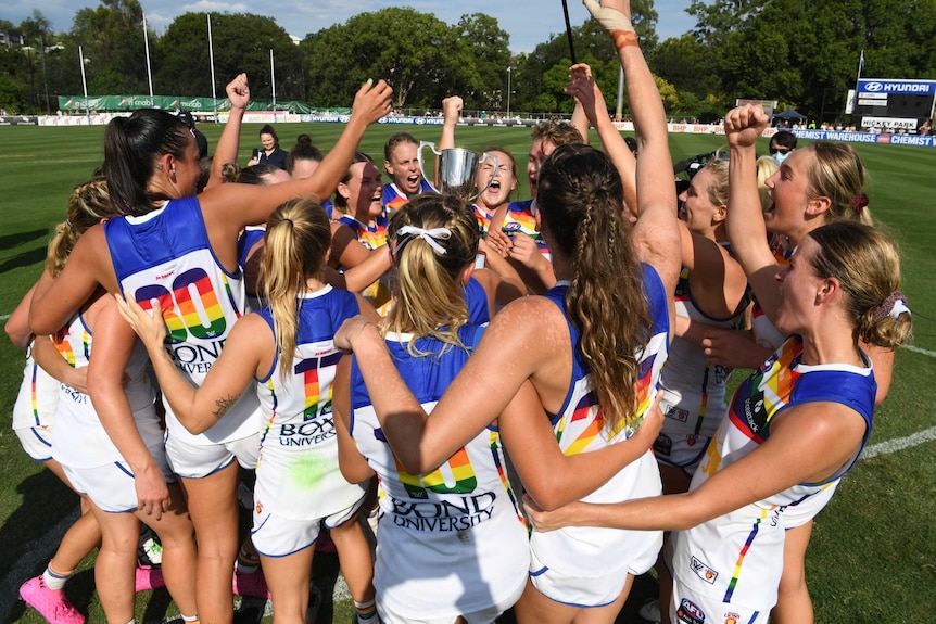 Brisbane Lions players celebrate in a huddle, while holding the Q-Clash trophy in the centre