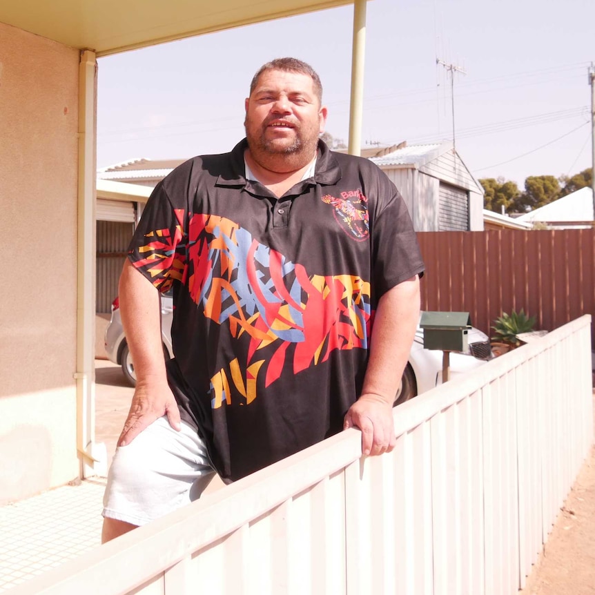 A man with a colourful T-shirt stands behind the front fence of a house on a residential street.