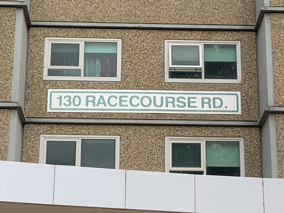 A sign reads '130 Racecourse Road' on the side of a high-rise public housing tower.