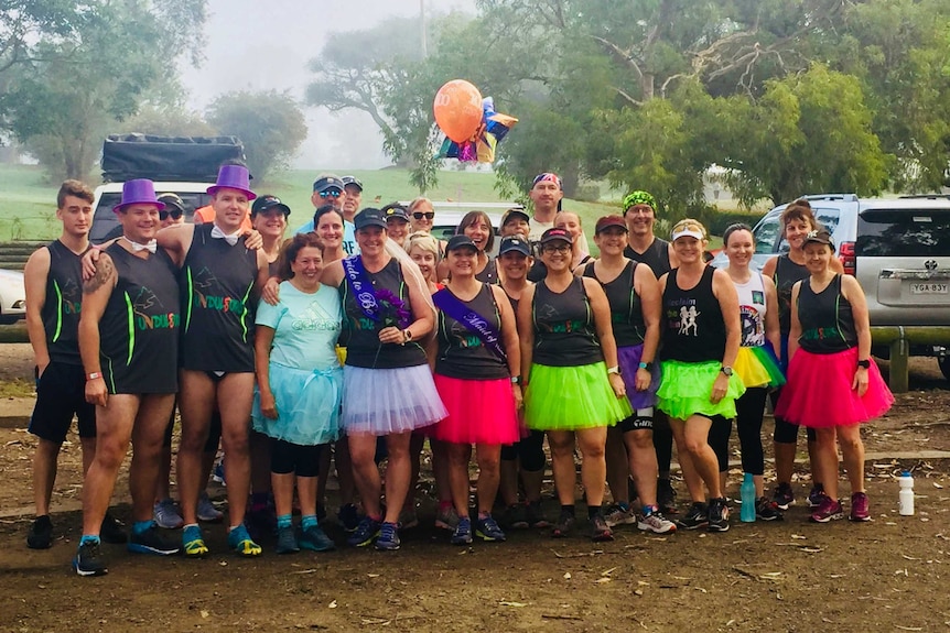 A group of people in running gear, accessorised with colourful tutus and novelty hats pose together at a park.