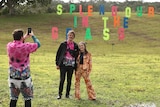 Two people stand in a field with large letters sitting on a hill behind them