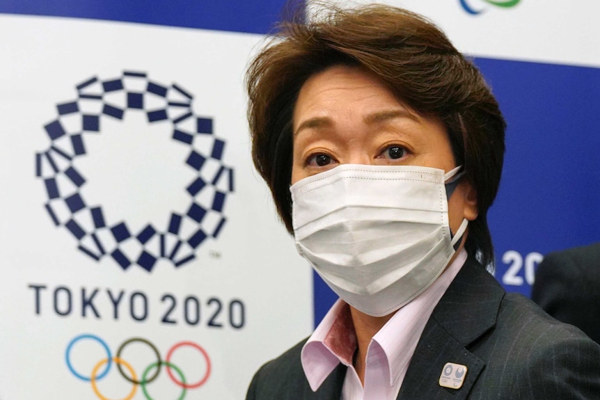 A mask-wearing Japanese Olympics organising executive stands in front of a Tokyo 2020 logo.